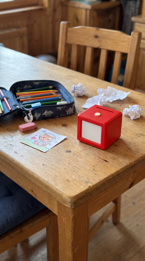 Fantasini steht mit Stiften, einem kleinen bunten Motiv und Bastelpapier auf dem Holztisch.